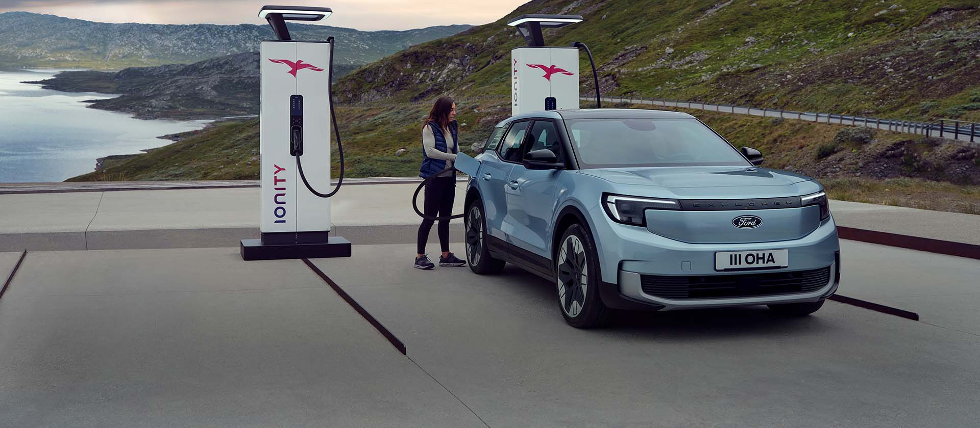 Egy nő kék, teljesen elektromos Ford Explorert csatlakoztat kábellel egy köztéri töltőállomáshoz. A jármű hegyek és víz közelében parkol.