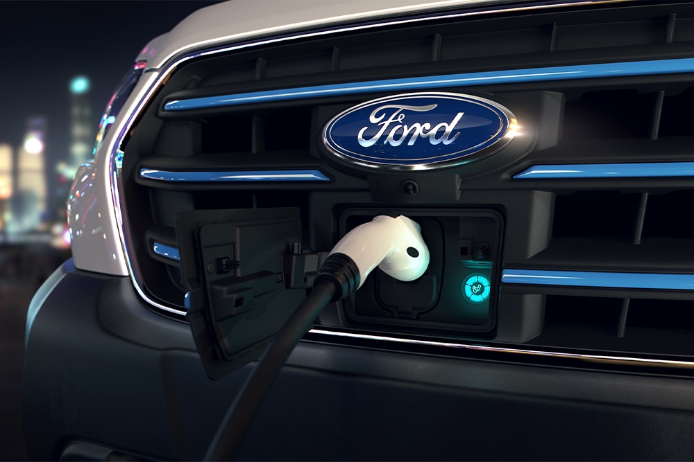Közelkép egy Ford E-Transit furgon töltőpontjáról és első homlokfalról a Ford logóval.