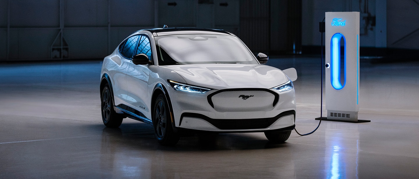 Egy garázsban áll egy fehér Ford Mustang Mach-E, és egy kék színben fénylő töltőállomáshoz csatlakozik.