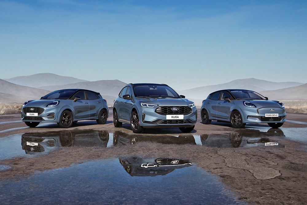 Egy parkoló Ford Puma, Ford Kuga és Ford Puma Gen-E modellt látunk egy esőáztatta úton, hegyekkel a háttérben.