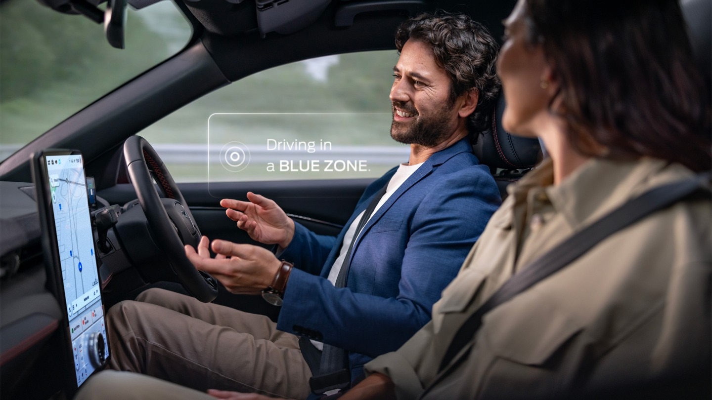 Egy férfi és egy nő kéz nélkül vezetnek egy Mustang Mach-E-t a Ford BlueCruise segítségével. Egy értesítés azt mondja: "Vezetés a BLUE ZONE-ban”. Egy férfi és egy nő kéz nélkül vezetnek egy Mustang Mach-E-t a Ford BlueCruise segítségével. Egy értesítés azt mondja: "Vezetés a BLUE ZONE-ban”.