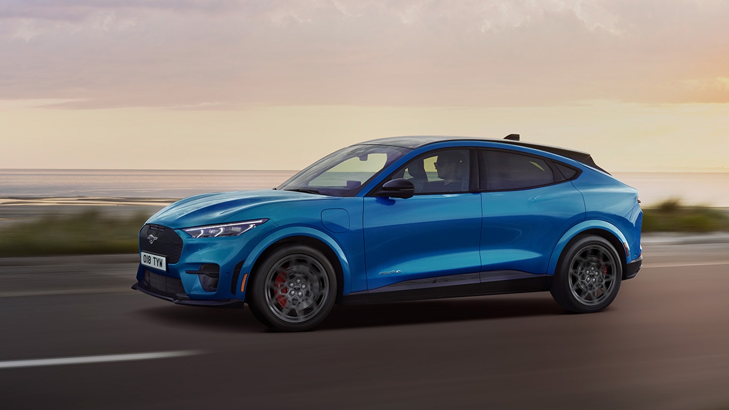 Kék színű elektromos Ford Mustang Mach-E oldalnézete alkonyatkor egy tengerparti úton.