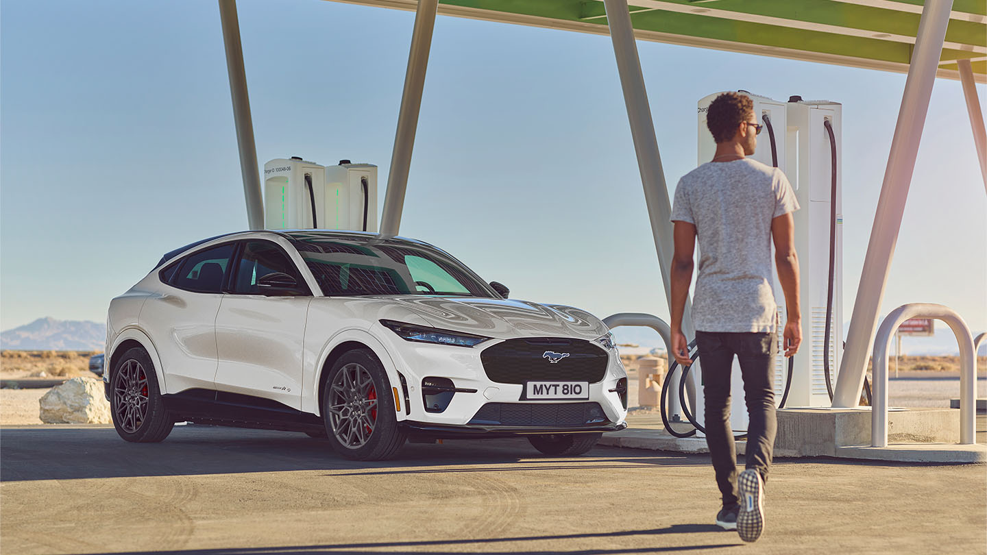 Köztéri töltőállomáson parkoló fehér Ford Mustang Mach-E.  Fehér pólót viselő férfi megy az autó felé.