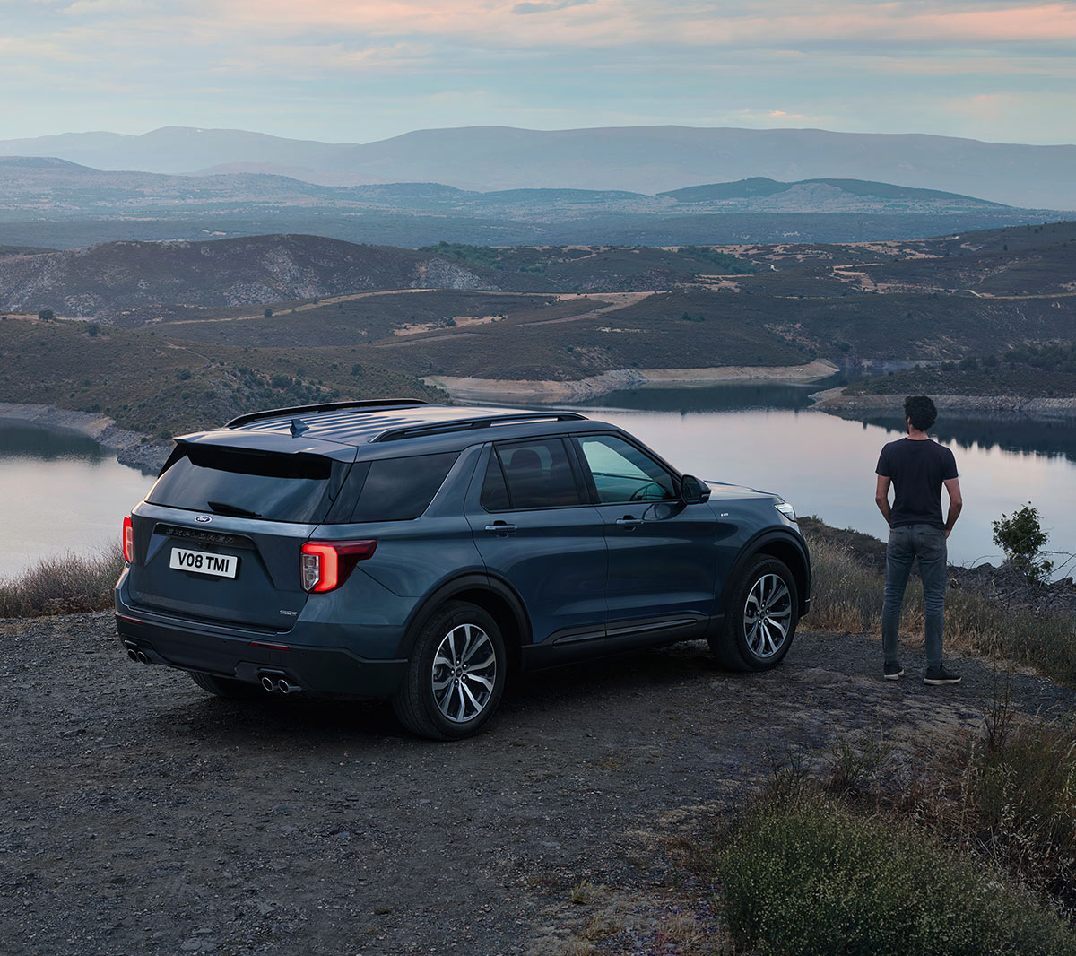 Ford Explorer parked in hilly area above lakes with person standing next to it overlooking landscape