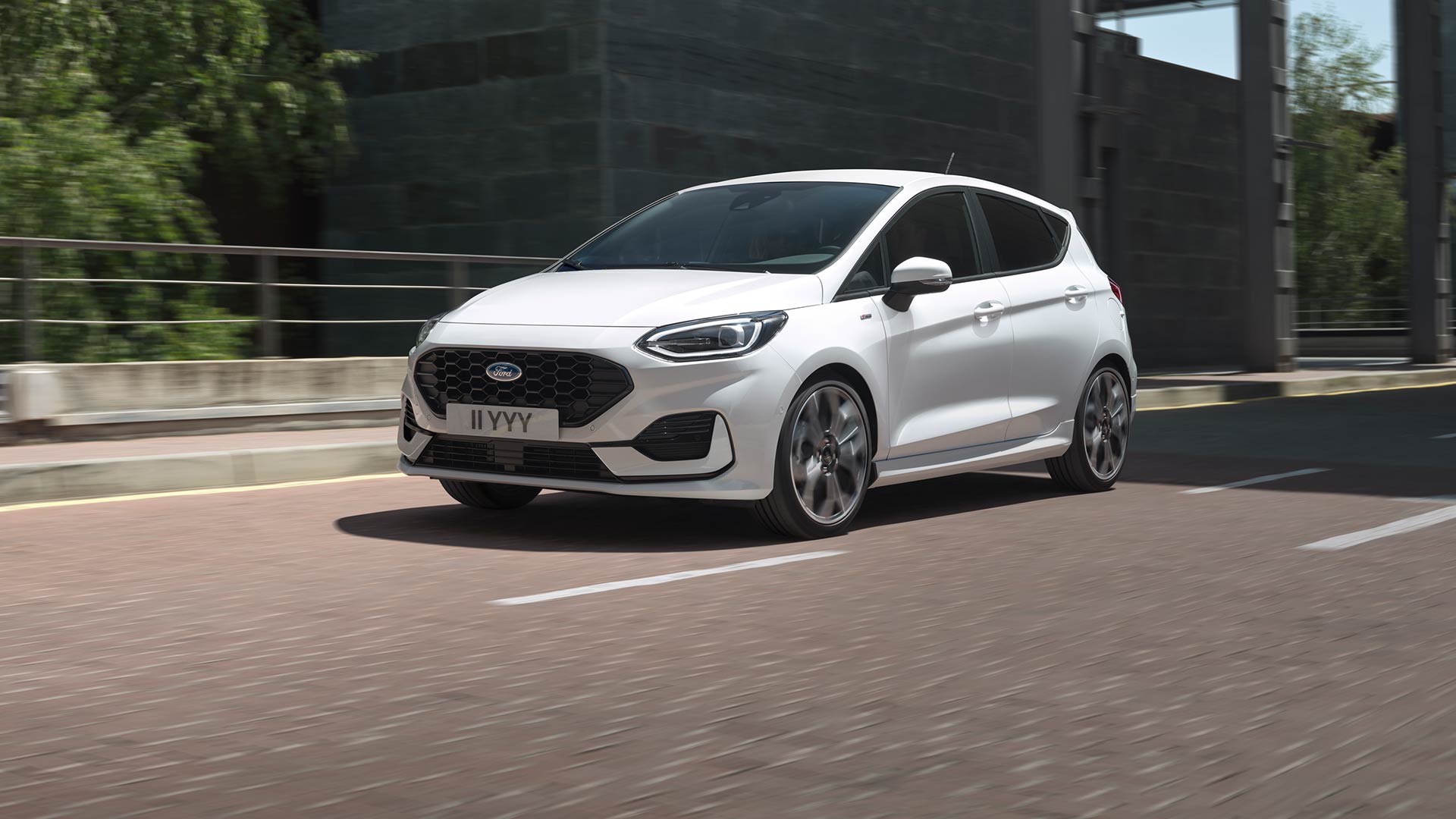 Egy fehér Ford Fiesta városi úton halad, a háttérben zöld növényzet és épületek láthatók.