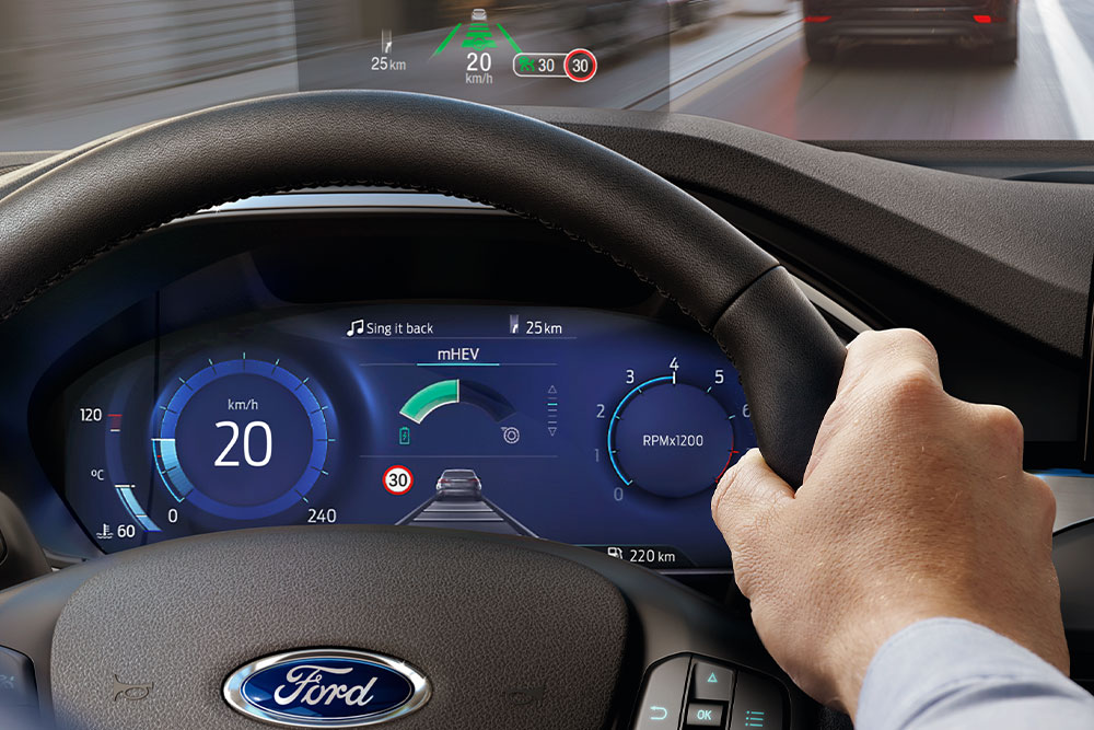 Egy kéz fogja a kormánykereket, a műszerfal fölött a Head-Up Display látható