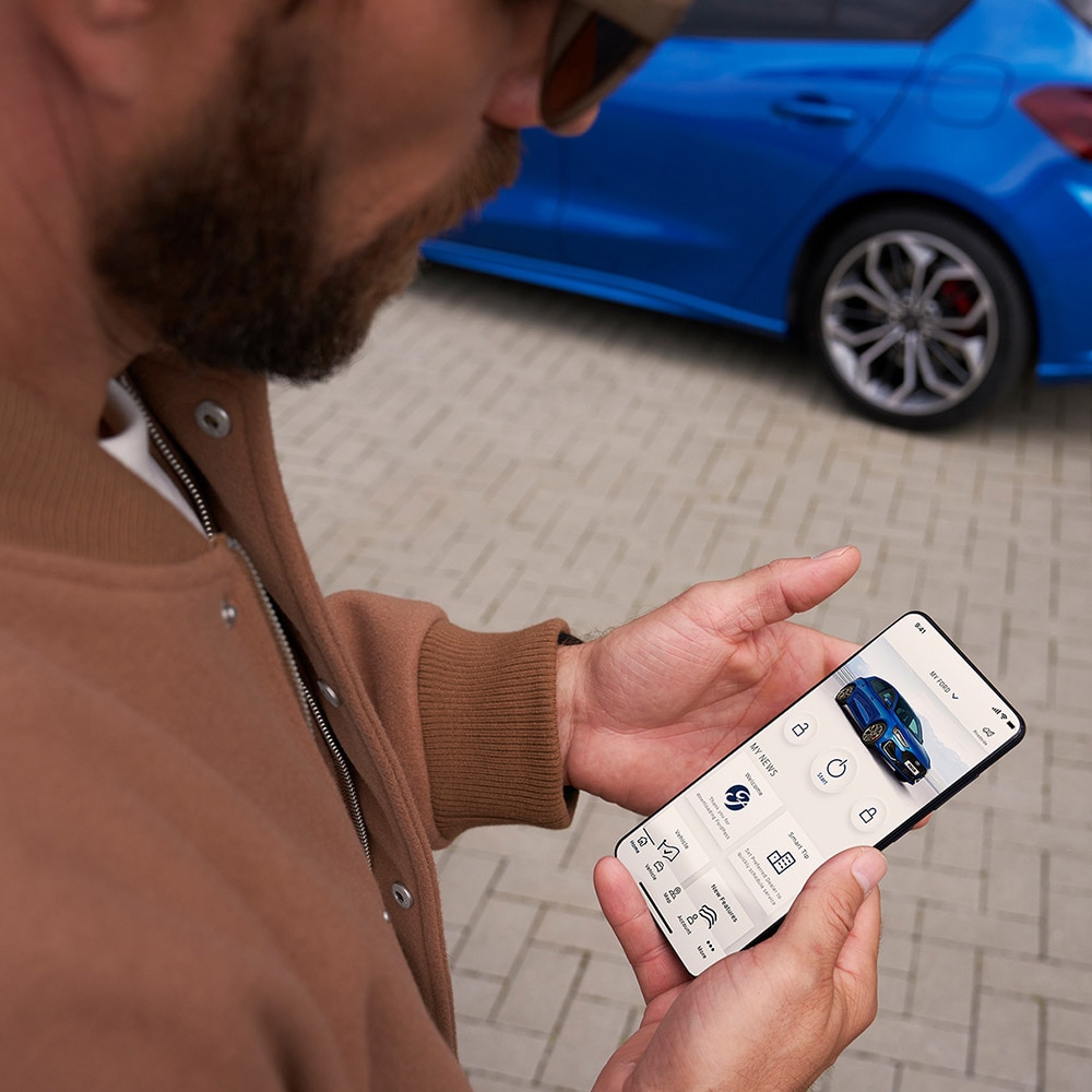 FordPass™ alkalmazást futtató okostelefont kézben tartó személy, a háttérben egy kék Ford Focus.