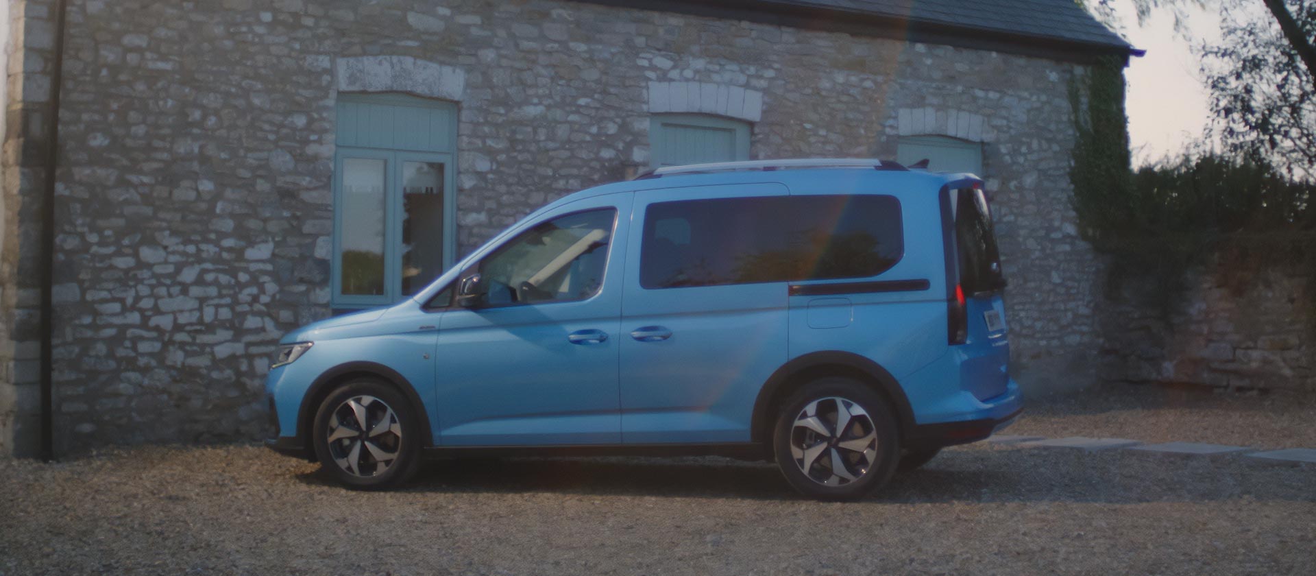 Egy kék Ford Tourneo oldalnézete, amely egy kőépület előtt, kavicsos felületen parkol.