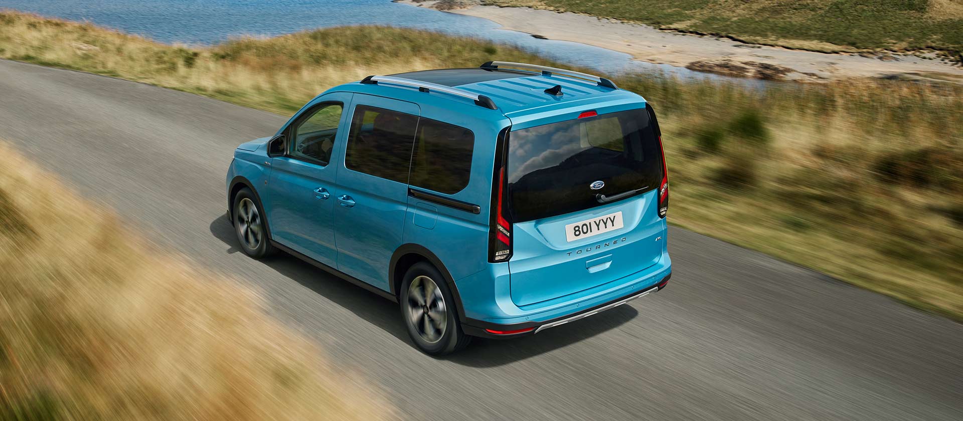 Kék Ford Tourneo hátsó 3/4-es nézete, amint egy kanyargós úton halad, látható tetőcsomagtartókkal.