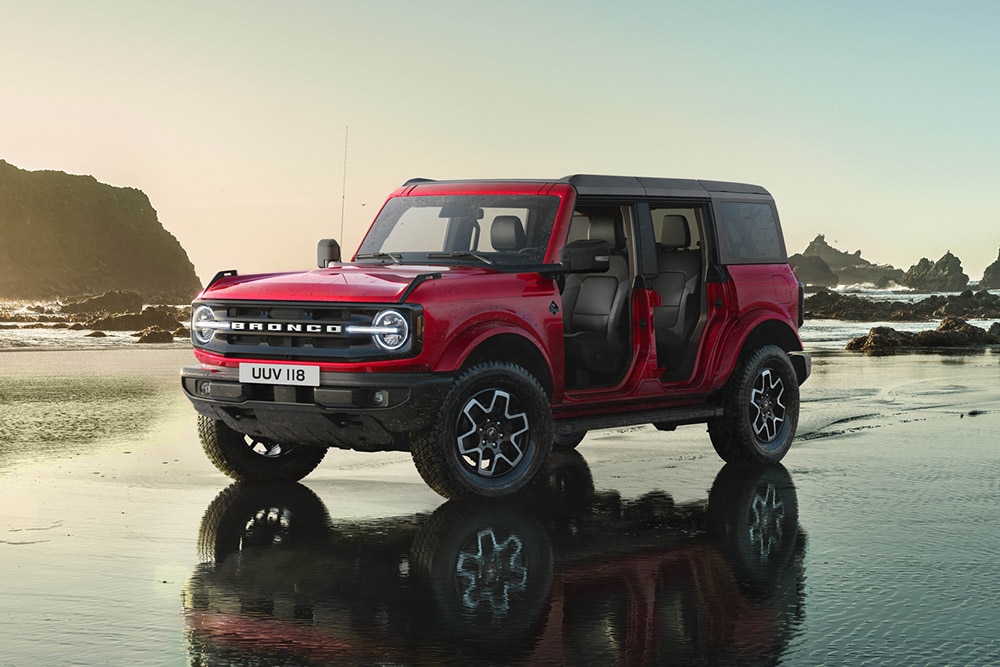 Egy piros Ford Bronco oldalnézete, ajtók nélkül, nedves homokos parton parkolva.