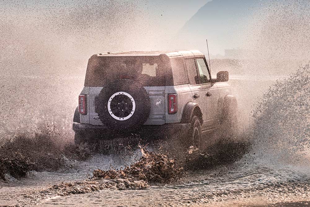 Ezüstszínű Ford Bronco hátulnézetből, amint a tengerpart átszelése közben felcsapja a vizet.