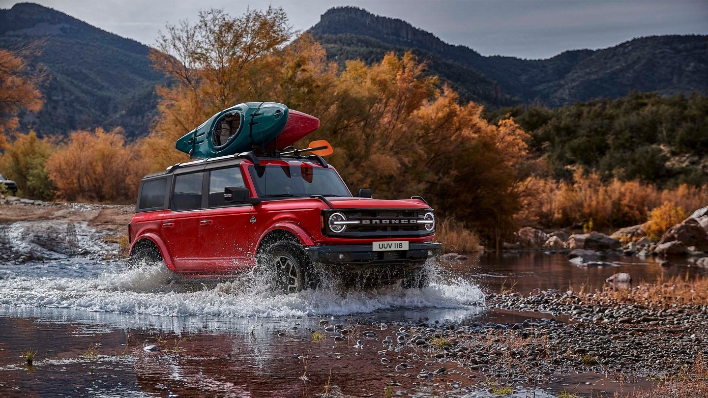 Ford Bronco driving through a small lake in the mountains with kayaks attached to the roof