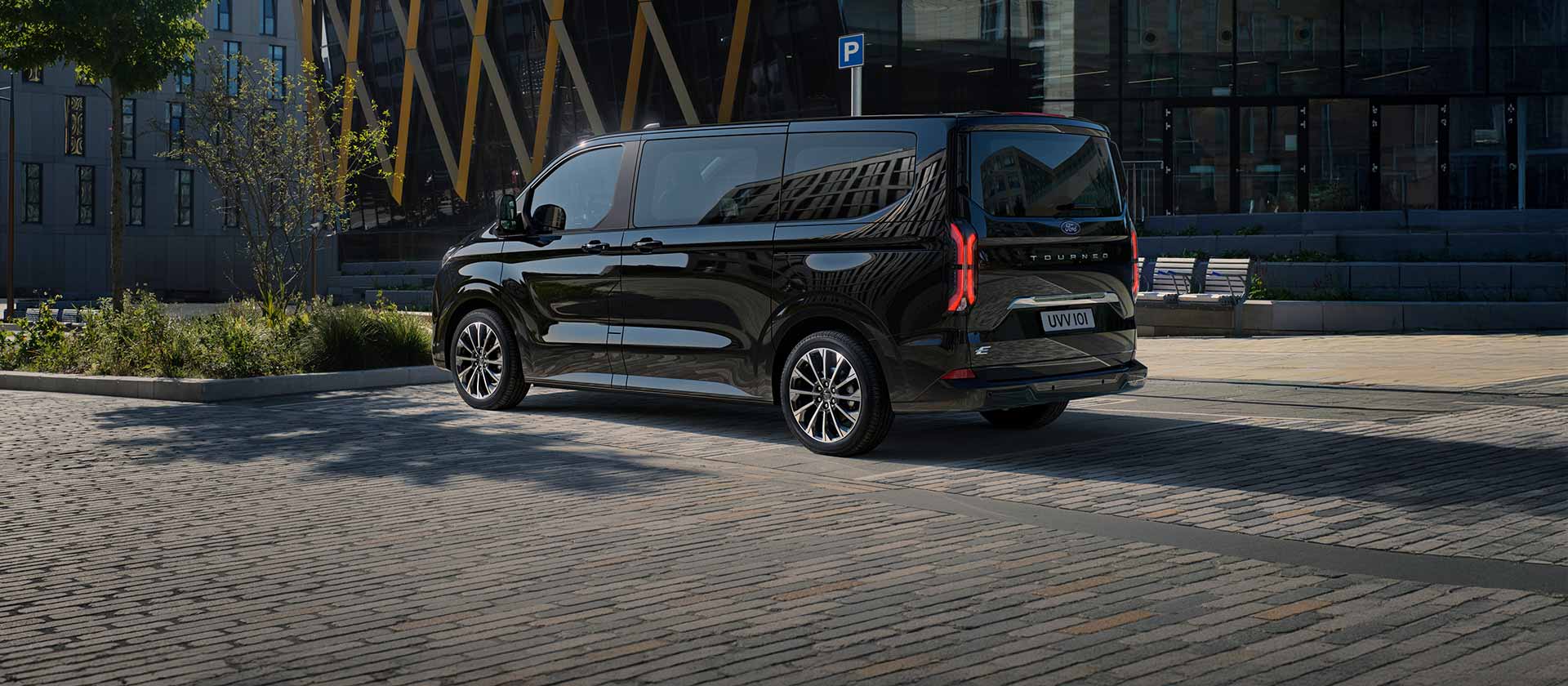 Fekete Ford E-Tourneo Custom oldal- és hátulnézetből, amint egy modern épület előtt parkol.