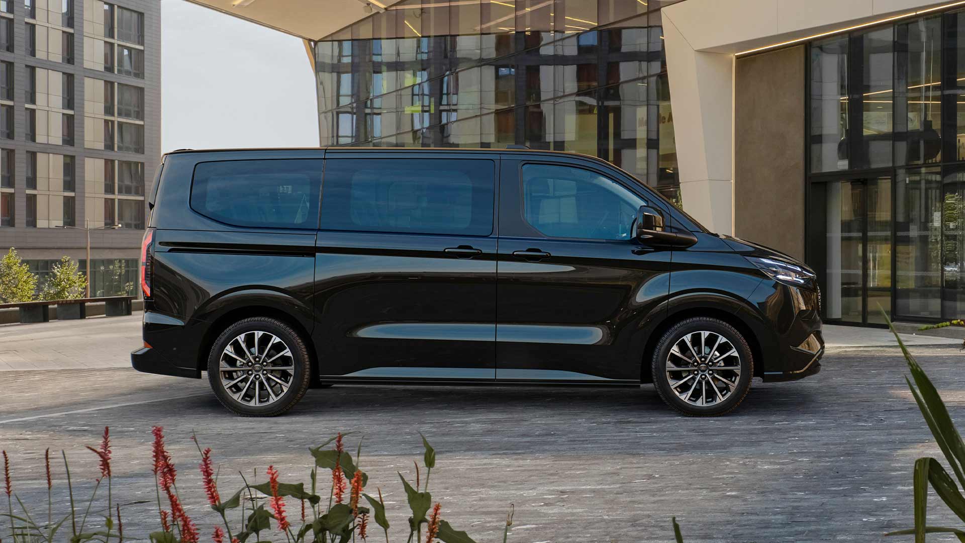 Egy fekete Ford E-Tourneo Custom egy modern épület előtt parkol.
