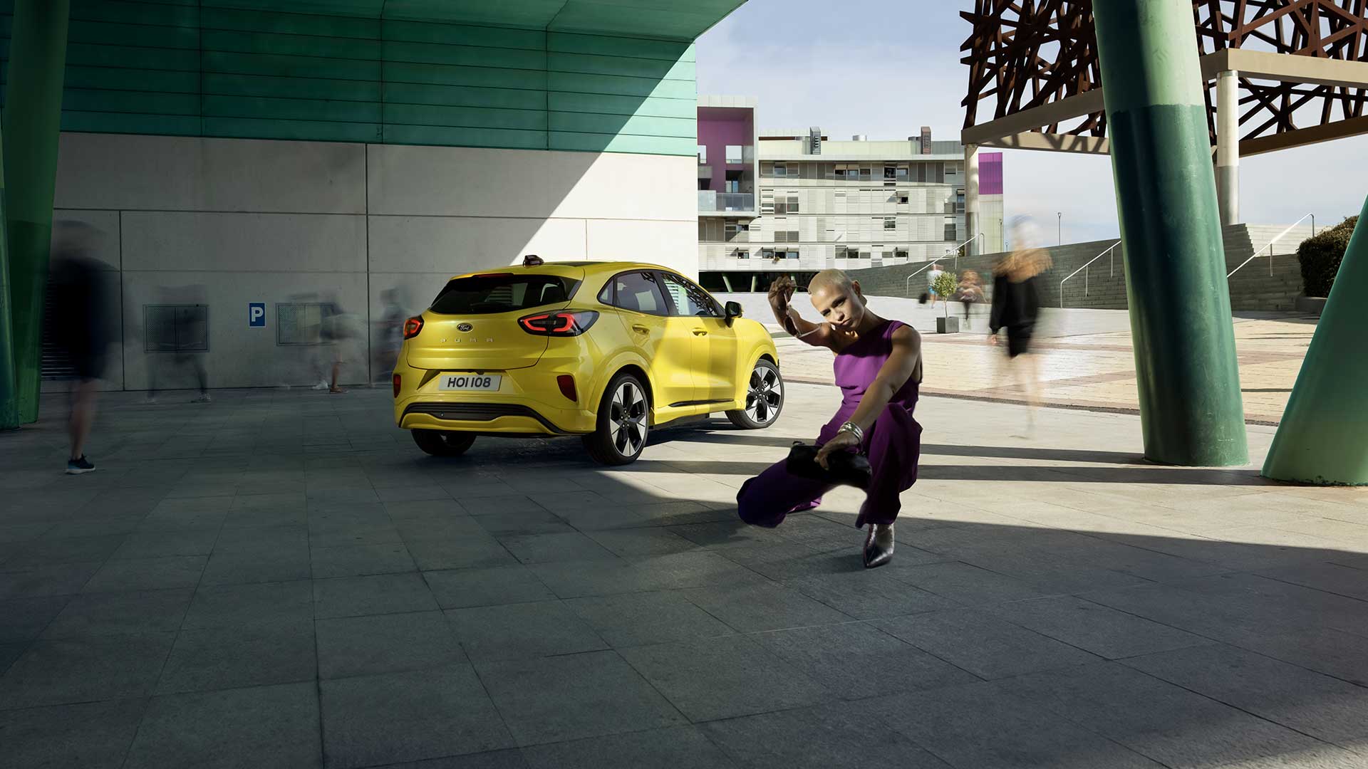 Híd alatt parkoló sárga, elektromos Ford Puma Gen-E® jármű hátulnézetből, egy lila ruhás személy mellett.