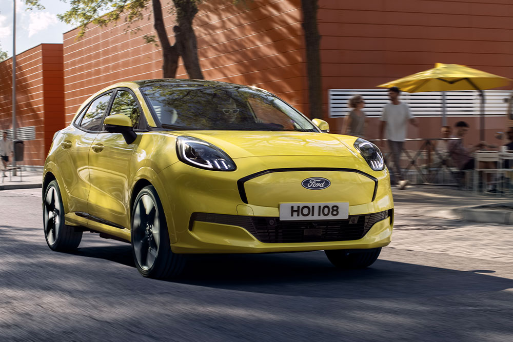 Városi úton haladó sárga, elektromos Ford Puma Gen-E® jármű elölnézetből.