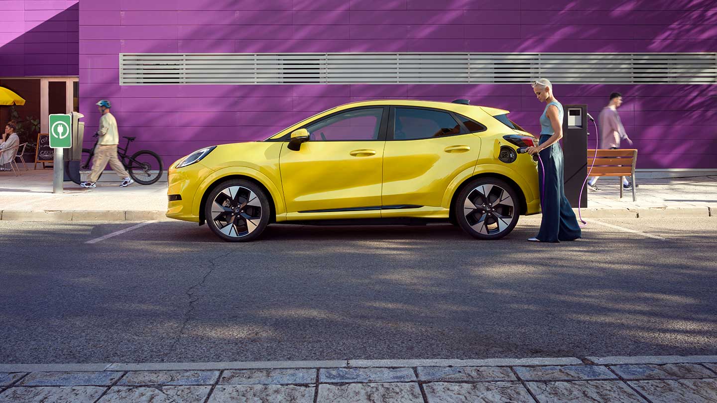 Egy parkolóhelyi köztéri töltőállomáshoz csatlakoztatott sárga, elektromos Ford Puma Gen-E® modell oldalnézetből.