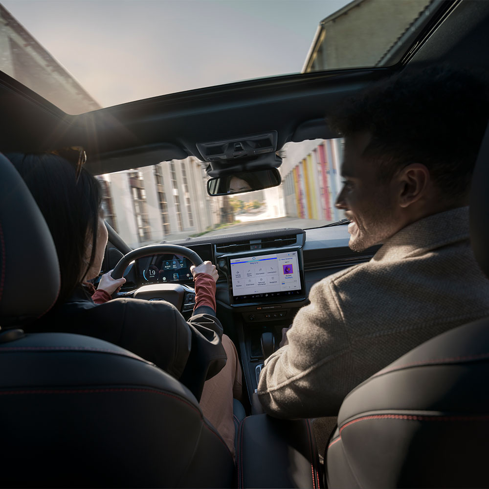 Egy vezető az utasával beszélget, az autó pedig egy napsütötte lakóövezeten halad át.