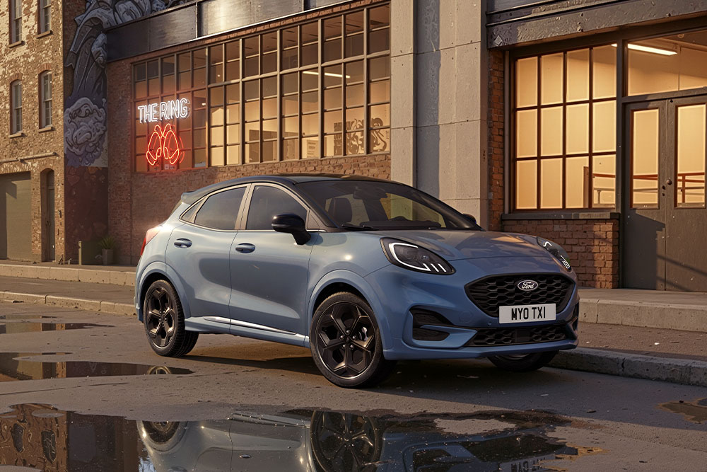 Egy leparkolt kék Ford Pumát látunk egy városi utcán naplemente idején, a víztócsákban az épületek tükröződnek.