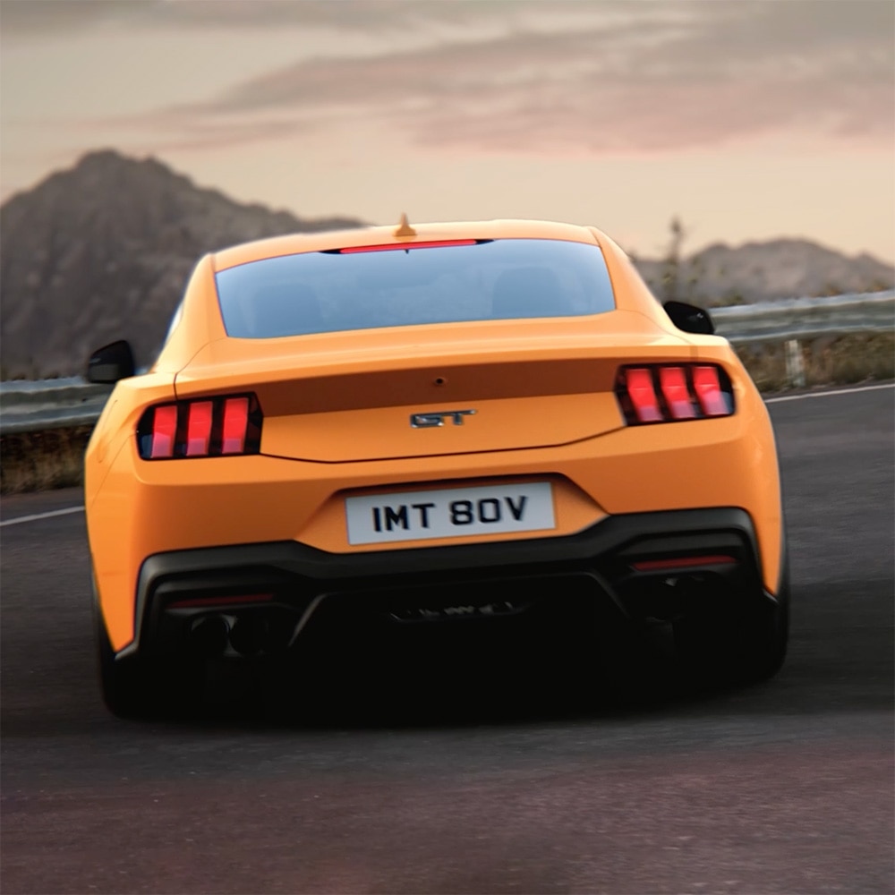 Egy Ford Mustang GT Fastback hátulnézete Orange Fury színben, amint szoros kanyart vesz egy vidéki úton