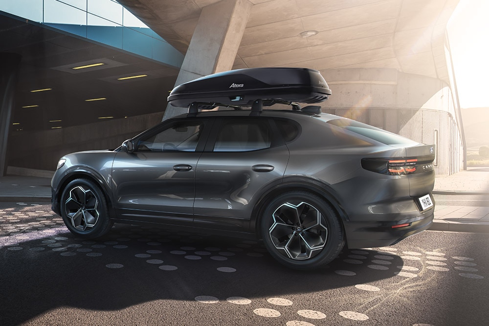 Sötét Ford Capri® tetőboxszal, oldalról 3/4-es nézetben, modern épületstruktúra alatt parkolva.