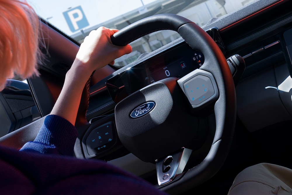 Közelkép egy nőről, aki egy leparkolt Ford Capriban a kormánykereket fogja. A digitális műszerfalat is látjuk.
