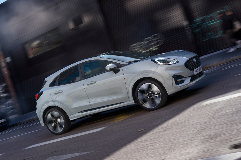 Nagyvárosi utcán halad egy ezüstszínű Ford Puma, a Ford városi autó termékcsalád egyik tagja.