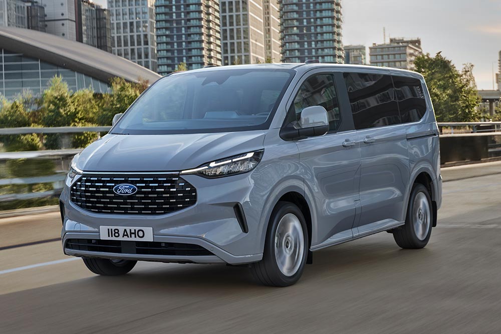 Egy ezüstszínű Ford Tourneo Custom egy városban halad az úton.