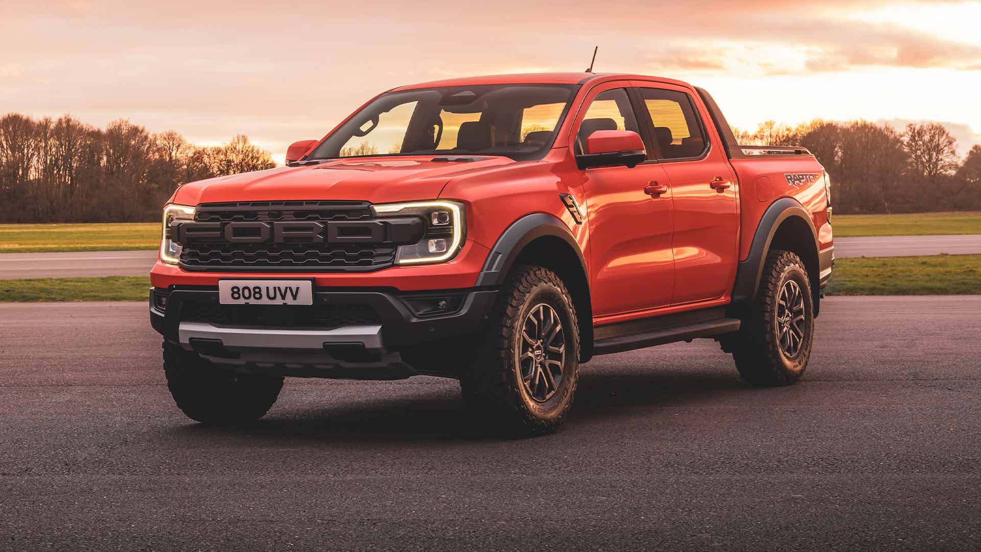 Egy Code Orange színű Ford Ranger Raptor egy úton parkol bekapcsolt fényszórókkal.