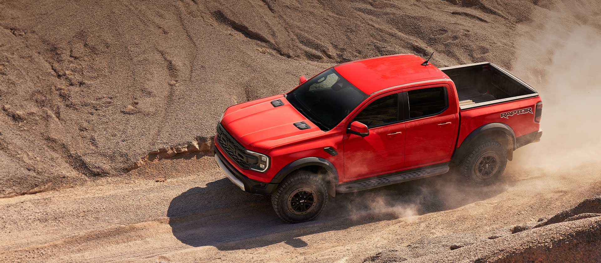 Egy Code Orange színű Ford Ranger Raptor hegymenetben, porfelhőben.