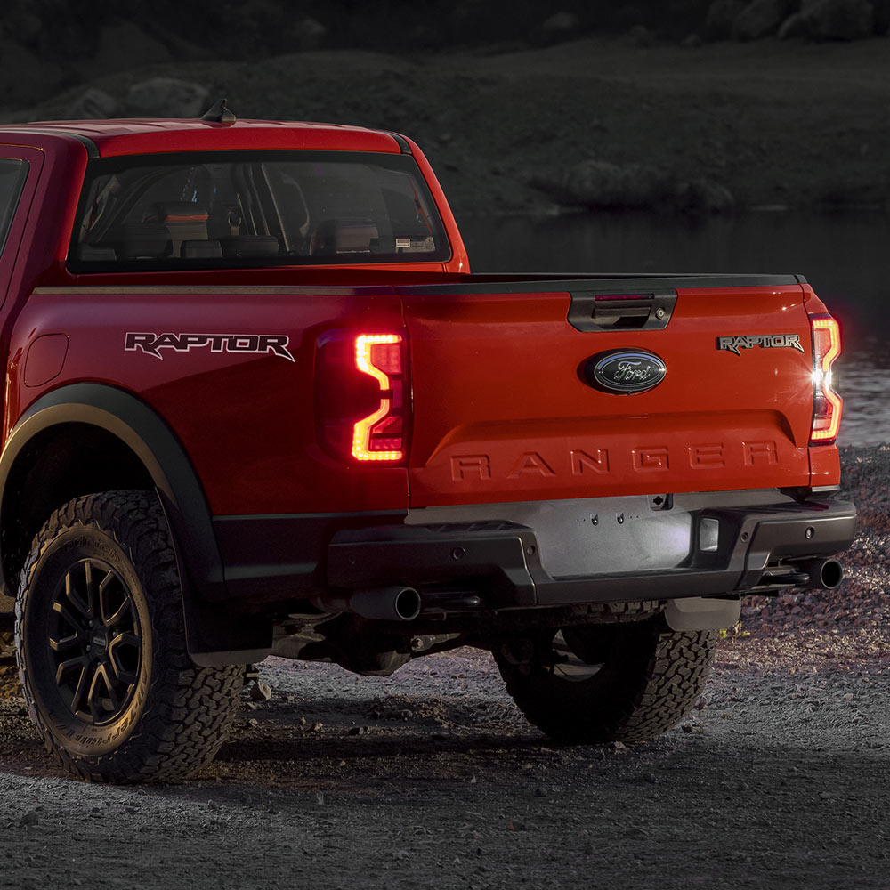 Egy Code Orange színű Ford Ranger Raptor bekapcsolt hátsó fényszórókkal hátulnézetből.