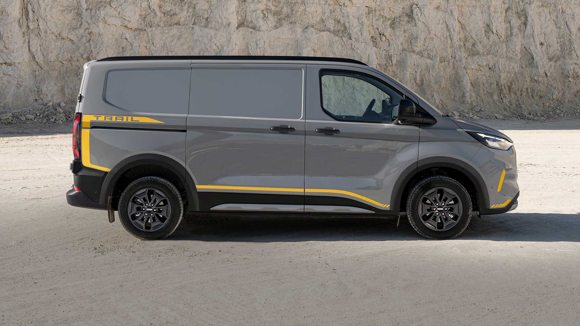 Egy szürke Ford Transit Custom Trail sárga dekorcsíkokkal, oldalnézetből.