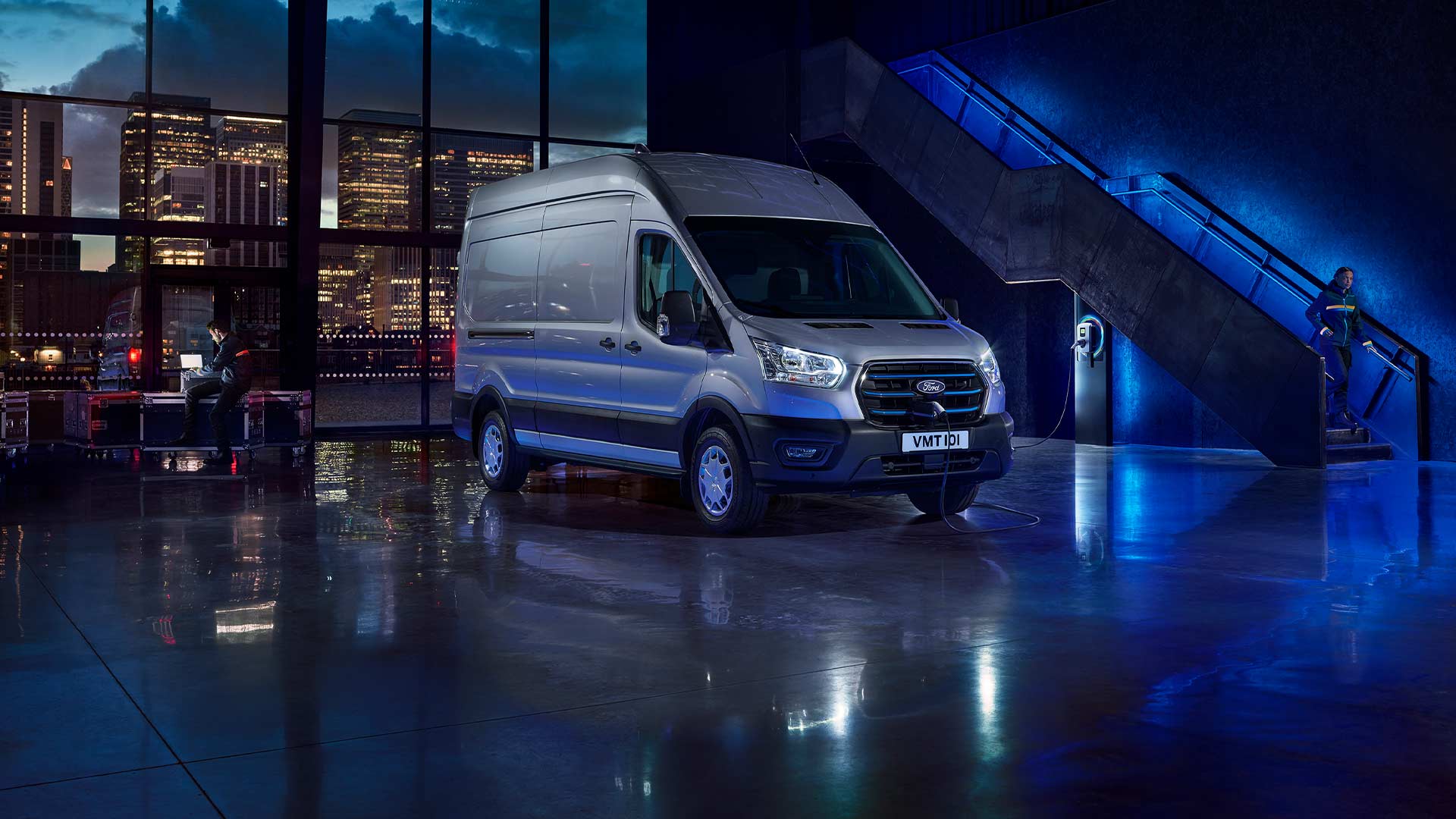 Egy ezüstszínű Ford E-Transit elektromos áruszállító parkol beltéren, nagy ablakokon át látható városi kilátással. 
