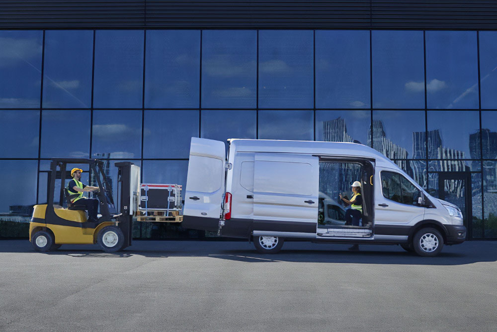 Két személy árut rakodik egy ezüstszínű Ford Transit áruszállítóba targoncával.