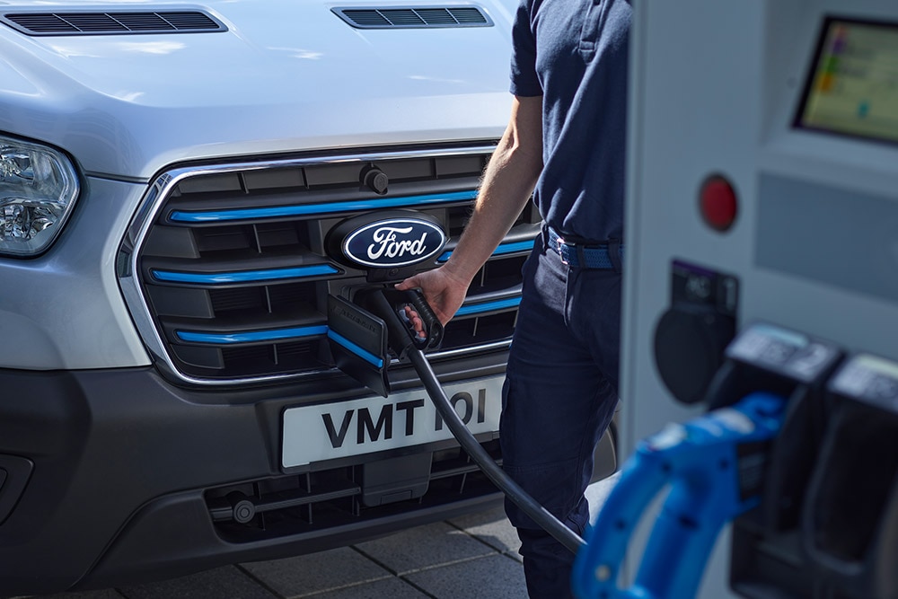 Egy személy egy elektromos töltőkábelt csatlakoztat egy ezüstszínű Ford E-Transit elektromos áruszállítóhoz.