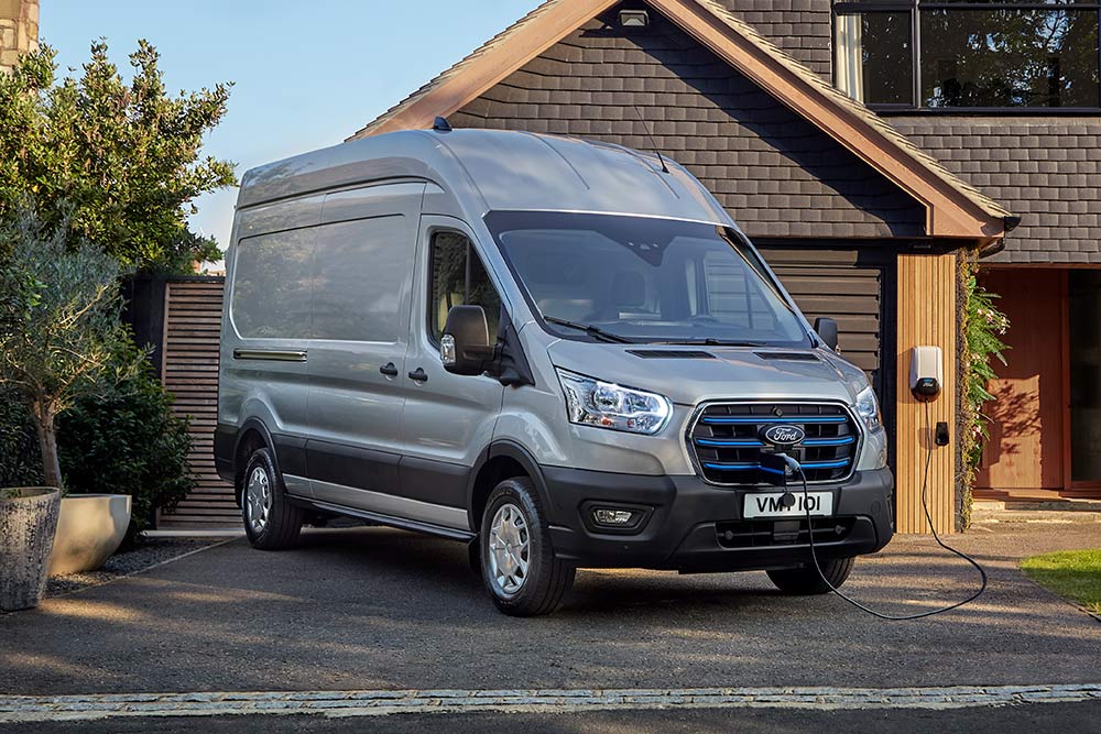 Egy ezüstszínű Ford E-Transit áruszállító egy ház melletti kocsifelhajtón parkol, töltőállomáshoz csatlakoztatva.