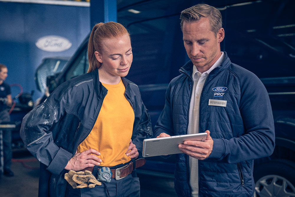 Két szerelő egy garázsban egy Ford E-Transit elektromos áruszállító előtt egy tabletet néz.