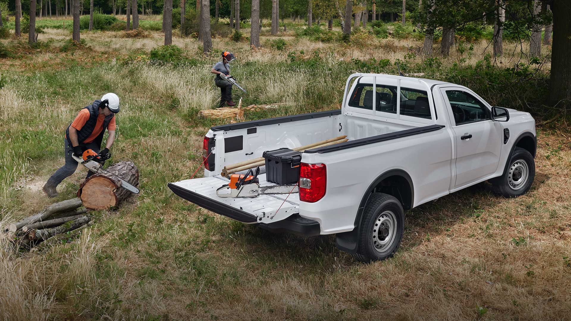 Normál kabinos, lehajtott hátfalú, fehér Ford Ranger parkol a füvön. Egy személy fát darabol láncfűrésszel a közelben.