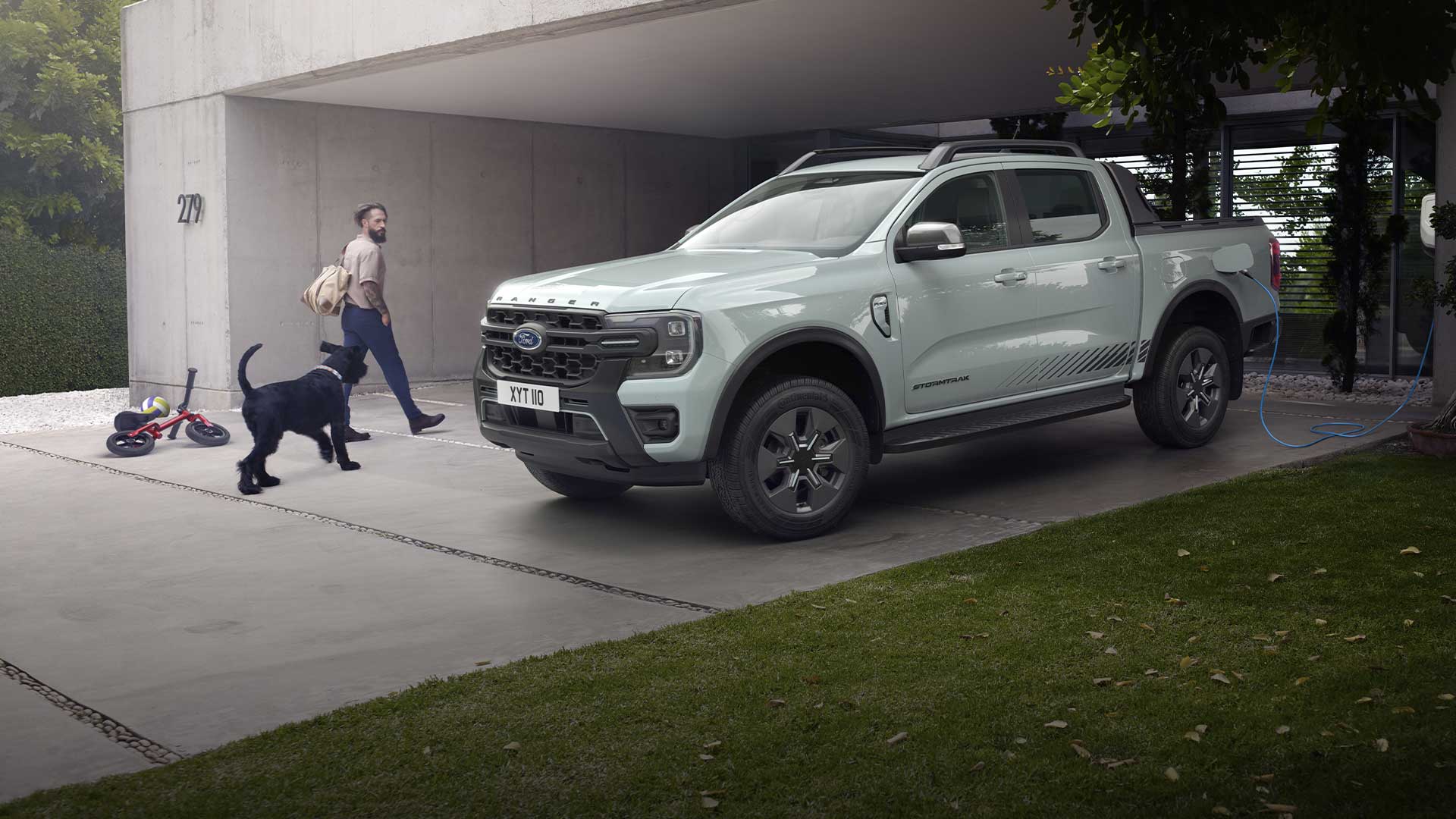 Egy kocsifelhajtón töltőre csatlakoztatott szürke Ford Ranger Plug-in Hybrid kisteherautó felé sétáló férfit és a kutyáját látjuk. 