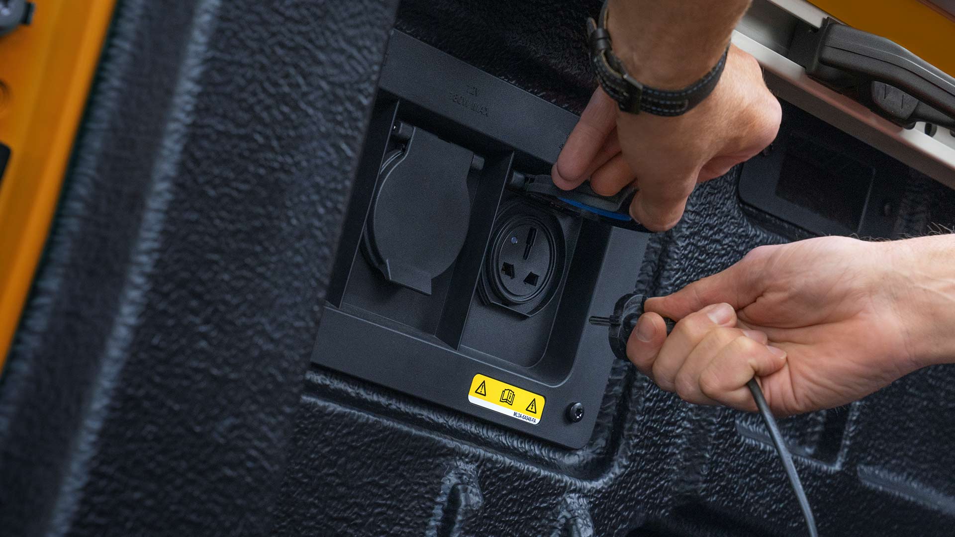 Közelképen látunk kezeket, amelyek a kábeleket a Ford Ranger platóján lévő elektromos aljzathoz csatlakoztatják.