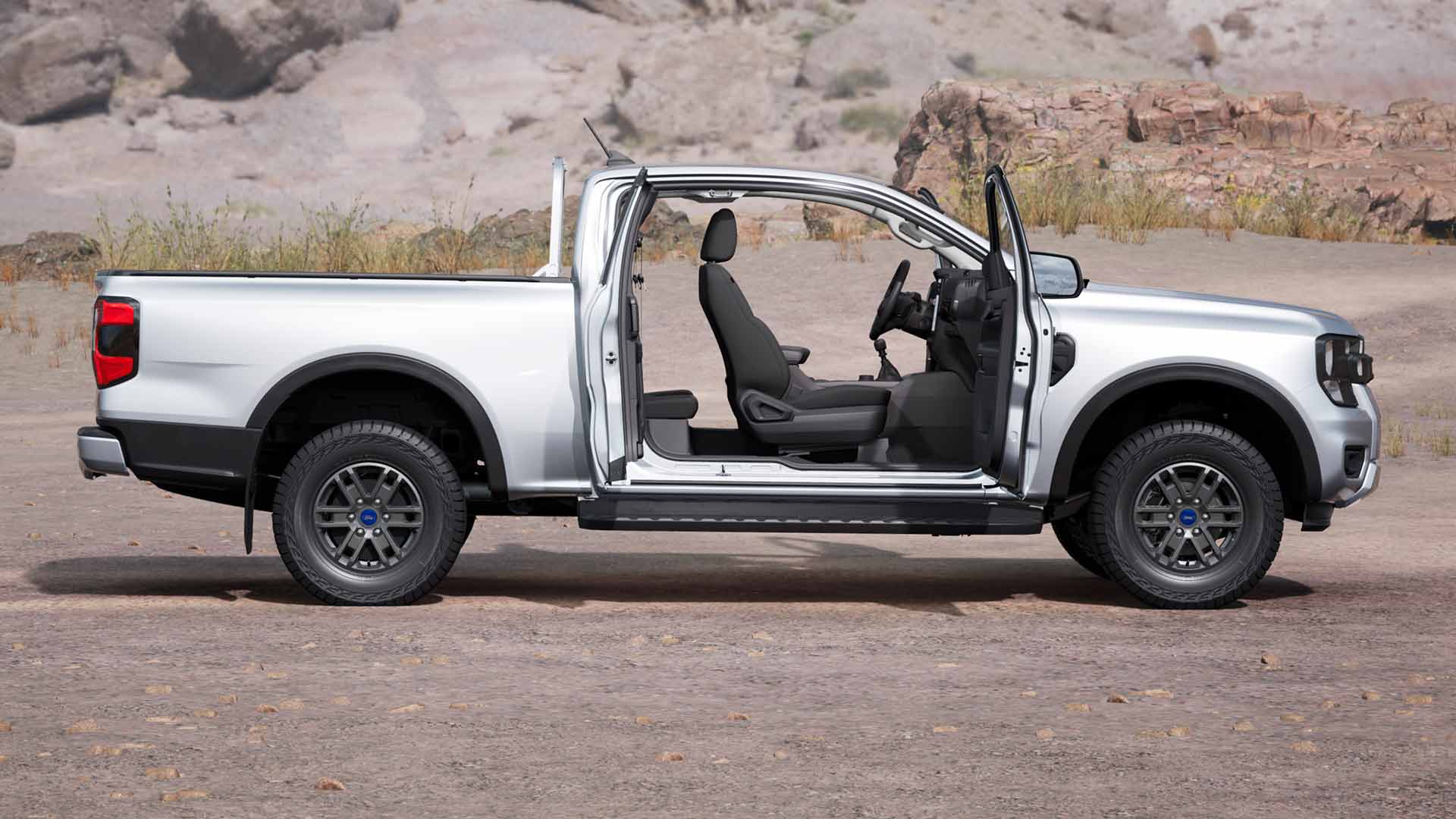 Oldalnézetből látunk egy ezüstszínű, nyitott ajtajú, poros terepen parkoló, nyújtottkabinos Ford Ranger XLT modellt.