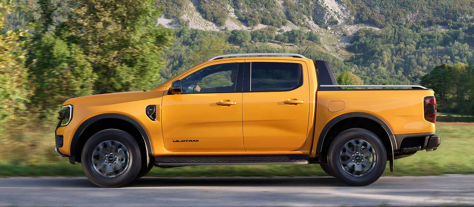 Oldalnézetből látunk egy sárga, duplakabinos Ford Ranger Wildtraket, amely egy hegyi út mellett parkol.