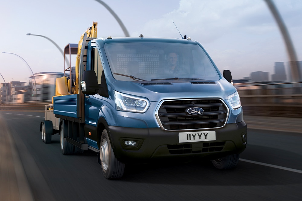 Ford Transit fülkés alváz elölnézetből, amely egy kotrógépet szállít, miközben városi úton halad.