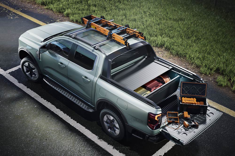 High-angle rear view of a grey Ford Ranger pickup truck, bed open with tools, and roof rack on a paved surface.