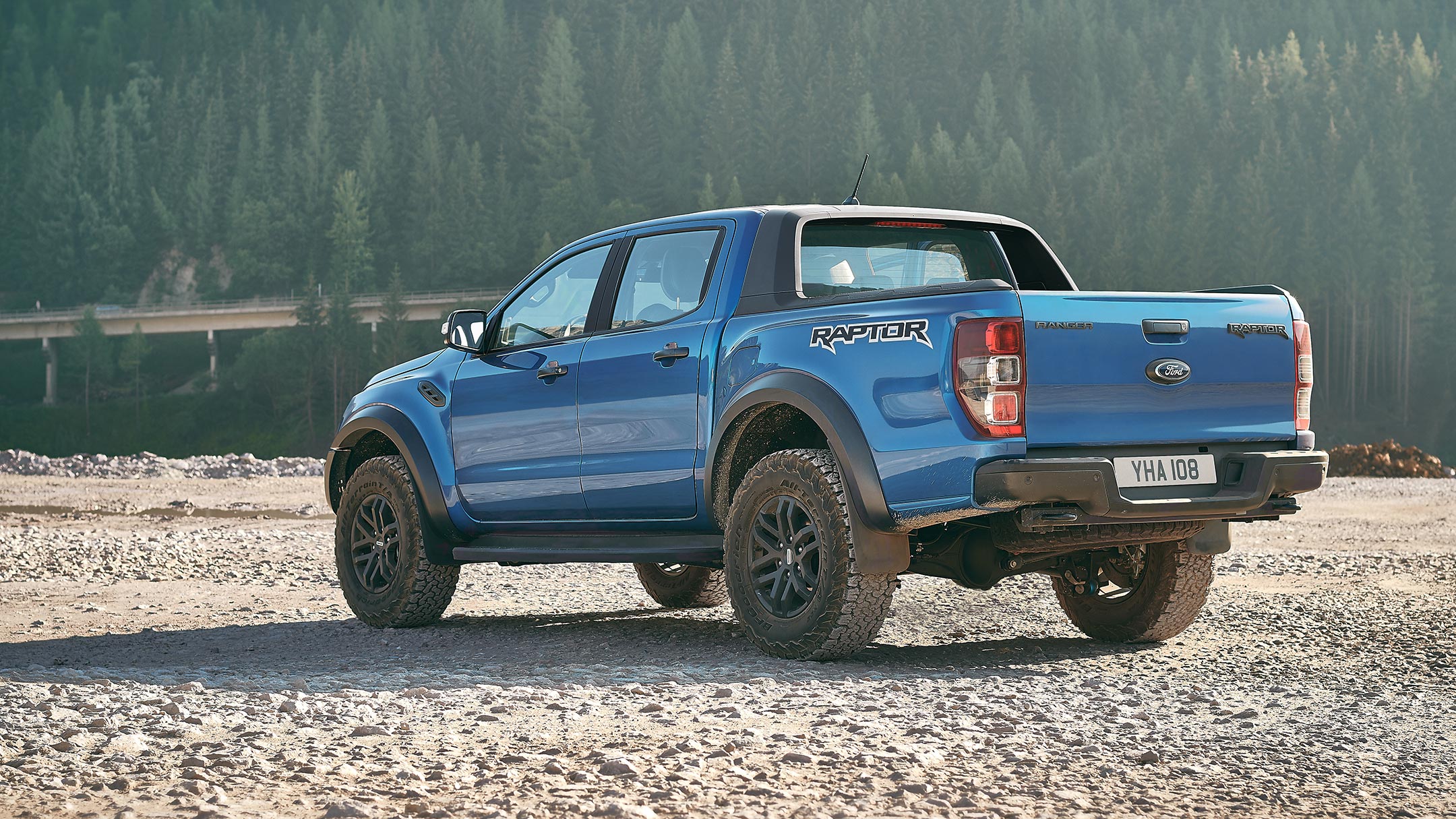 Kék Ford Ranger Raptor a terepen egy erdő mellett parkol, félig hátulnézetben