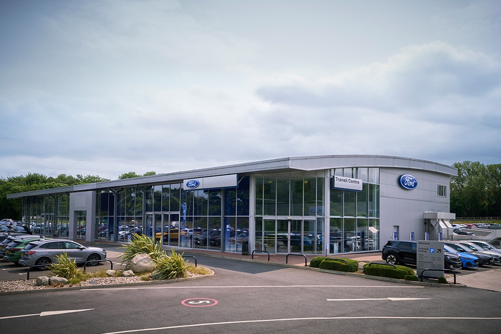 Kívülről látunk egy modern Ford márkakereskedést, amely előtt különböző Ford járművek parkolnak.