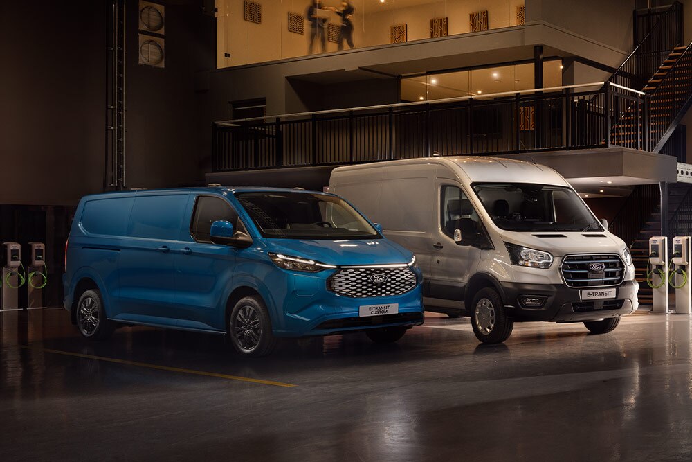 Egy kék Ford E-Transit Custom és egy ezüst Ford E-Transit töltődik egy parkolóban.