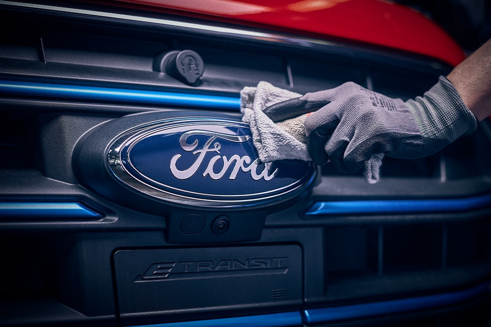  Kesztyűs kéz megérinti a Ford emblémát egy jármű első hűtőrácsán.