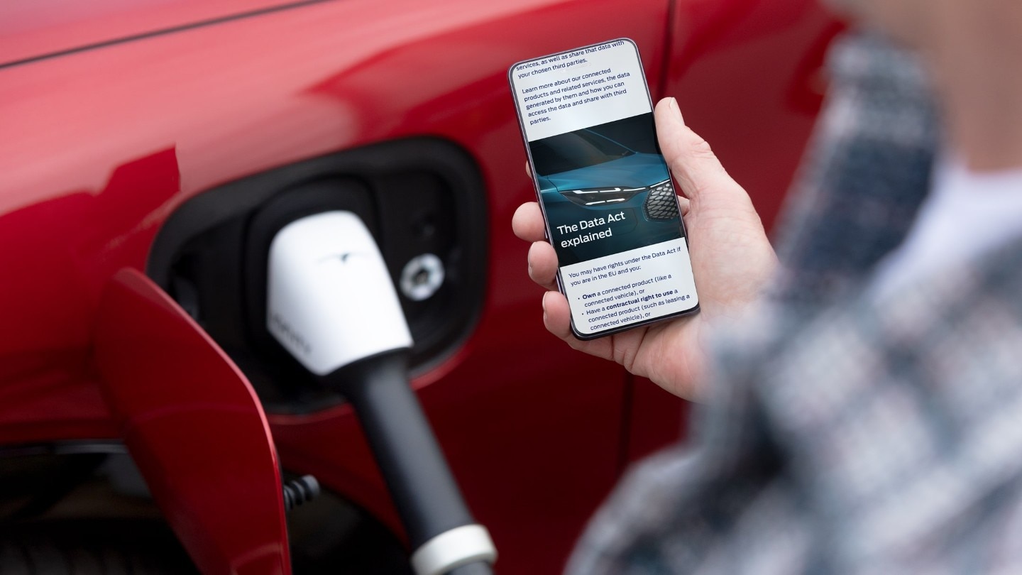 Egy személy az okostelefonjával van elfoglalva, a háttérben egy vörös színű elektromos jármű töltődik.